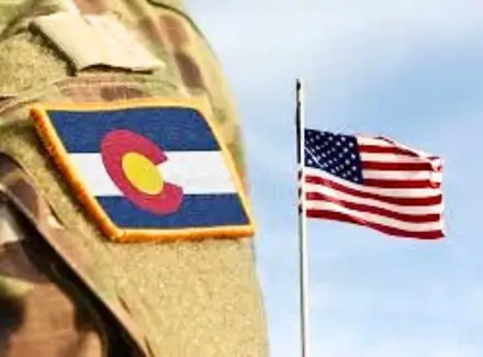 A military solider in uniform with Colorado state patch on his shoulder and USA flag in the background.
